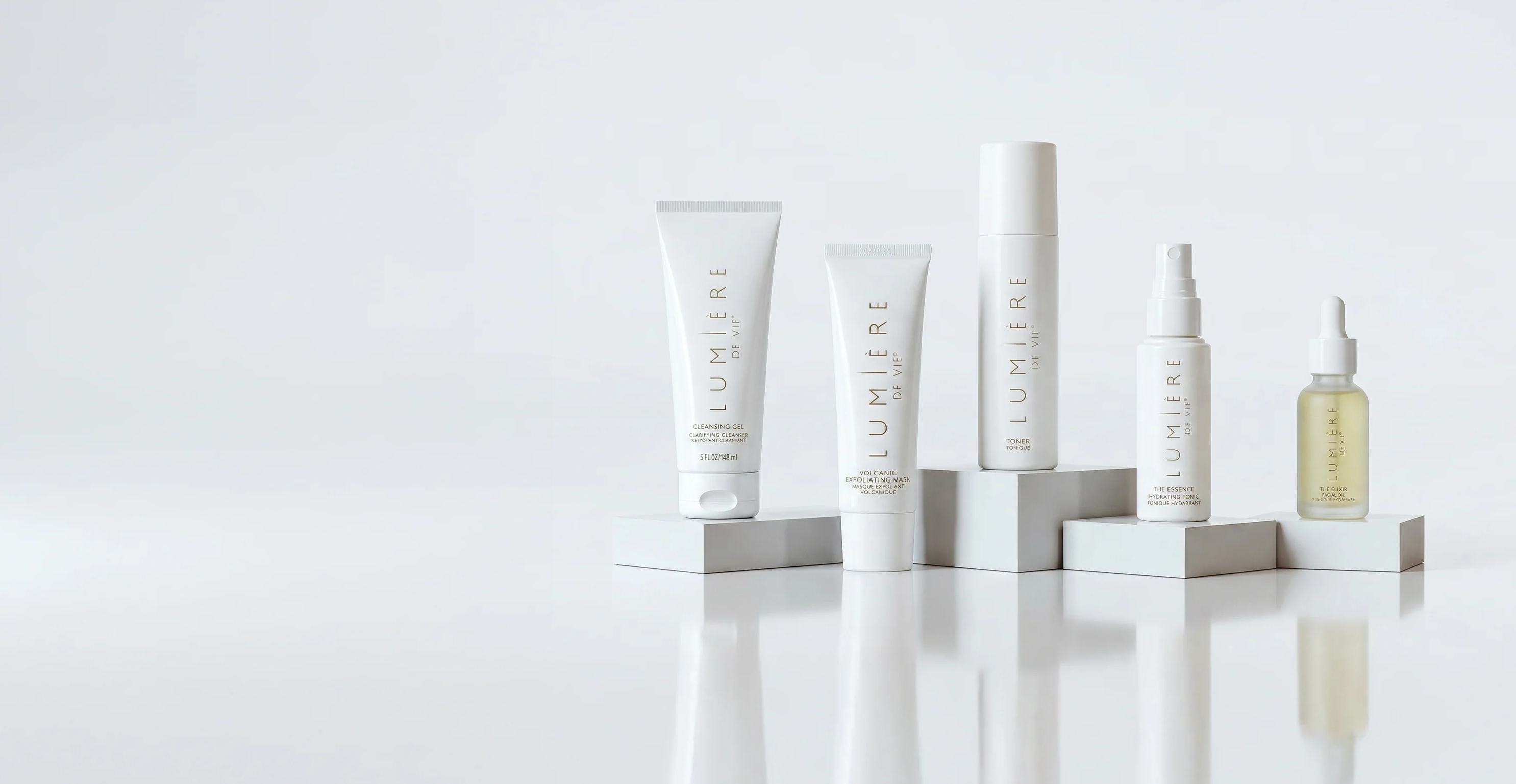 A set of five skincare products from the brand Bloomier, including tubes, a spray bottle, and a dropper bottle, arranged on minimalist white pedestals against a clean white background.