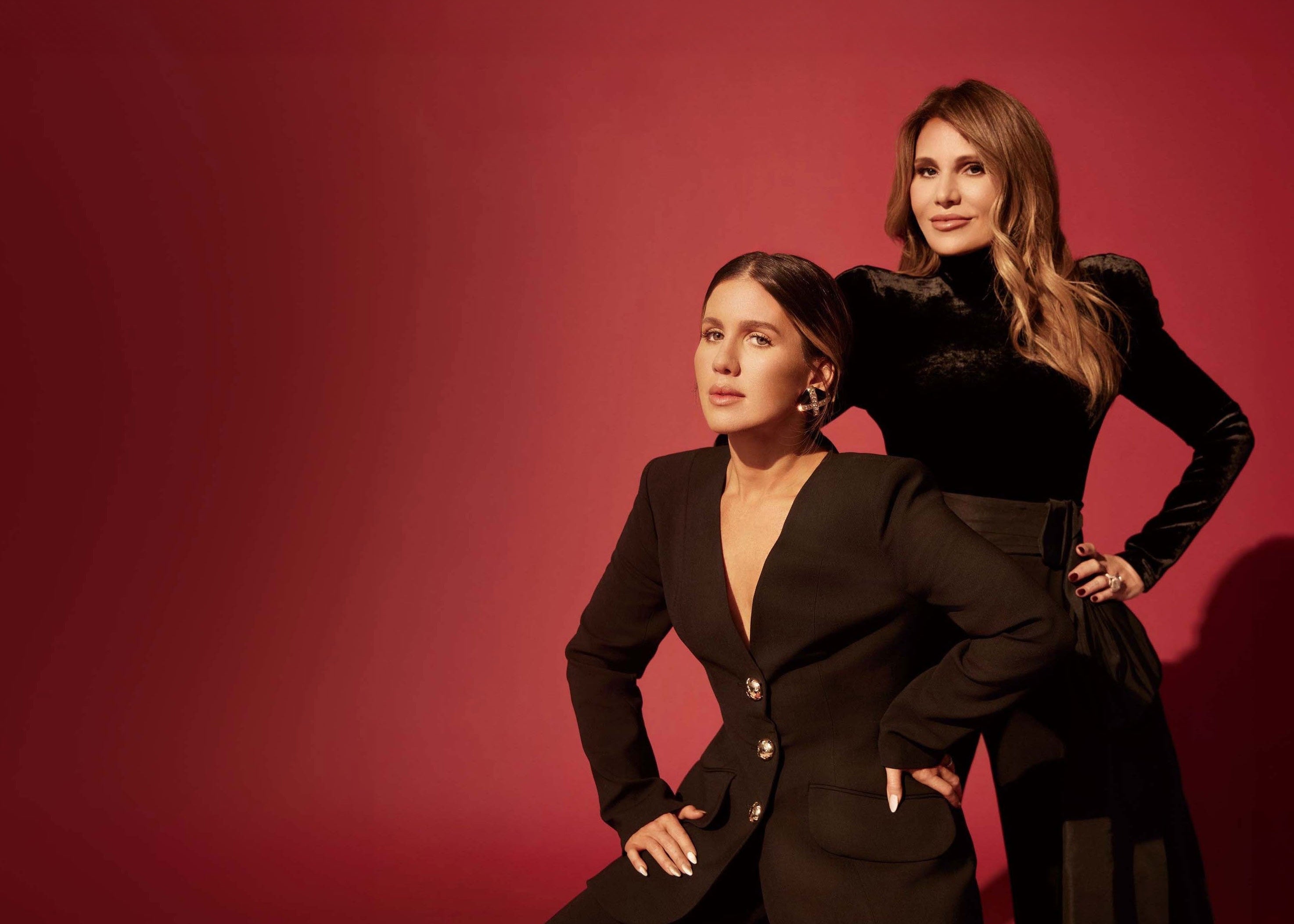 Two women in black outfits posing against a dark red studio backdrop with dramatic lighting and shadows.