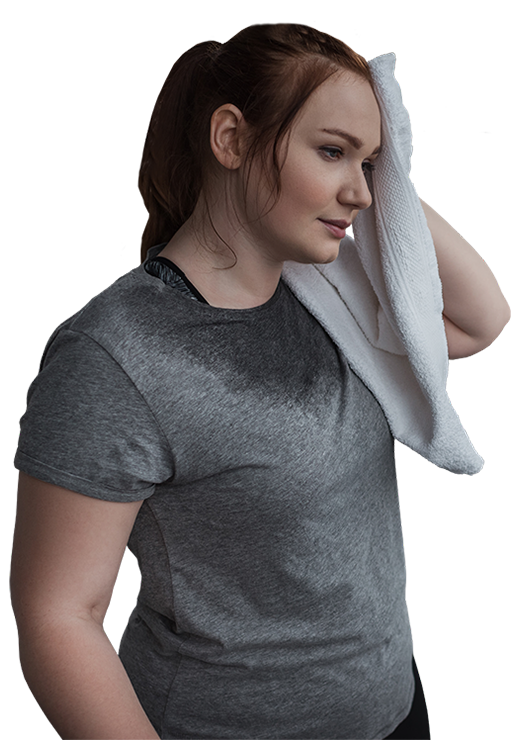 A woman with red hair in a ponytail wearing a dark gray heathered t-shirt with a wet stain on the front, holding a white towel to her forehead against a solid white background.