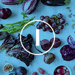 A close-up, top-down view of a blue surface scattered with dark purple grapes, red beets, blackberries, and green asparagus, overlaid with a white circular icon containing a vertical bar.
