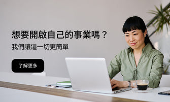 A woman with dark hair sits at a desk using a silver laptop, wearing a sage green blouse. A cup of coffee is on the table next to her.