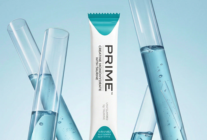 A single PRIME tooth powder stick pack stands upright in the center, flanked by several clear glass test tubes filled with light blue liquid against a pale blue background.
