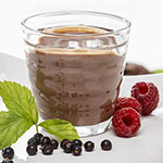 A clear glass cup filled with a chocolate brown smoothie or drink, shown on a white surface with raspberries, blackberries, and a green leaf around it.