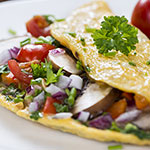A close-up of a prepared omelette or breakfast sandwich on a white plate, filled with vegetables including red tomatoes, white onions, and green parsley.