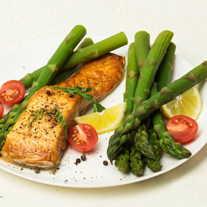 A plated meal featuring two pieces of cooked salmon with lemon slices, cherry tomatoes, and green asparagus on a white plate.