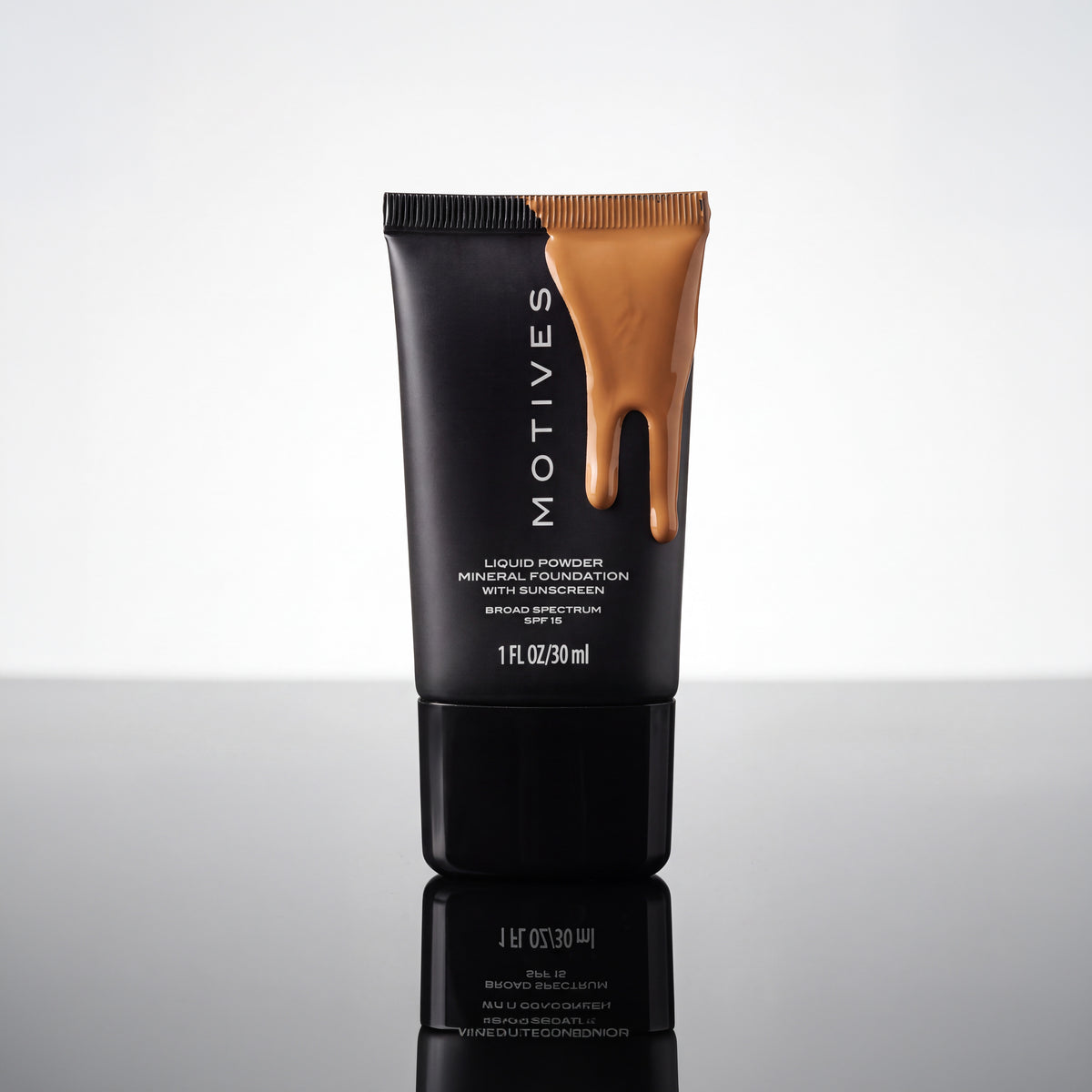 A black squeeze tube of MOTIVES Liquid Powder Foundation with a glossy brown liquid dripping from the top, set against a reflective light gray background.