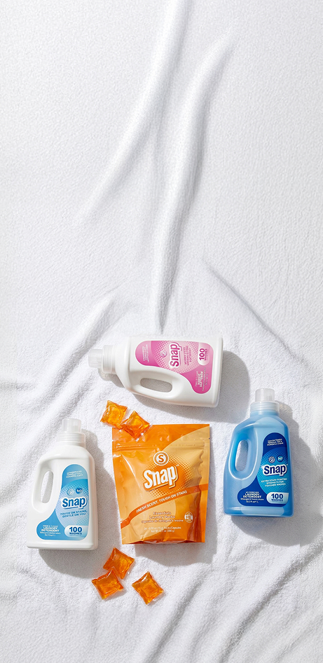Four Snap laundry detergent products arranged on a white textured fabric background: a white bottle with blue label, a white bottle with pink label, an orange bag, and a blue bottle, with small orange packets scattered nearby.