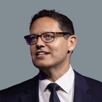 Man wearing glasses, a suit, and tie against a gray background