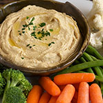 A bowl of thick beige hummus topped with a drizzle of oil and chopped green herbs, surrounded by fresh vegetables including broccoli, carrots, and cauliflower on a white background.