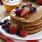 A stack of golden pancakes topped with fresh strawberries, blueberries, and syrup on a white plate, with a small bowl of honey in the background.