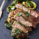 Sliced cooked beef with a green herb sauce on a dark plate, with a fork partially visible at the top left.