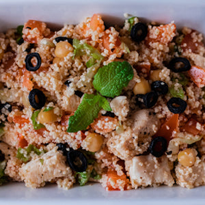 A close-up shot of a prepared couscous salad in a white bowl, featuring cooked couscous mixed with diced tomatoes, chickpeas, black olives, green herbs, and a mint leaf.