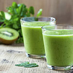 Two clear glasses filled with a bright green smoothie on a rustic wooden surface, with kiwi slices and green leaves blurred in the background.