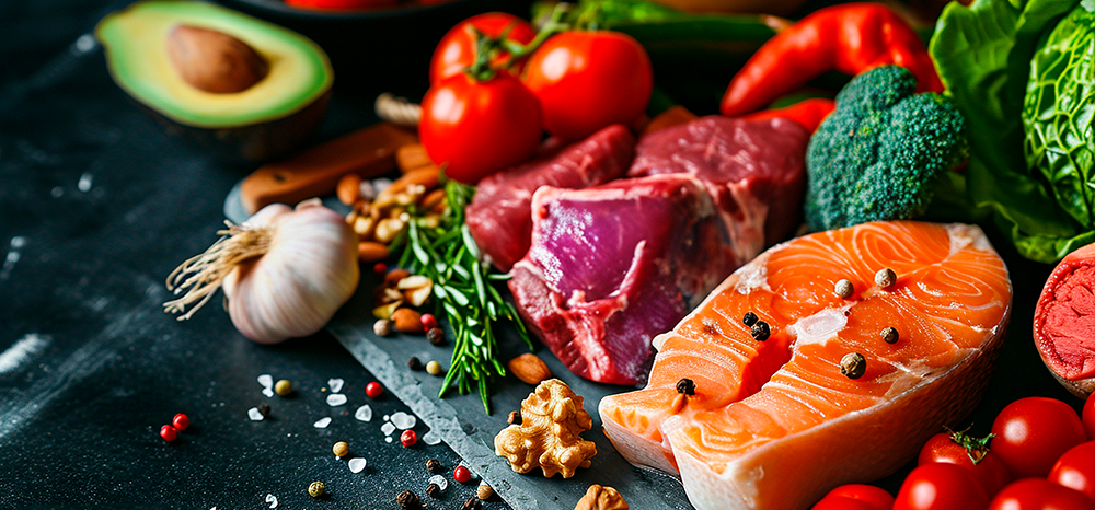A styled flat-lay of fresh raw foods on a dark slate board, including salmon fillets, red meat, broccoli, tomatoes, garlic, avocado, and walnuts.