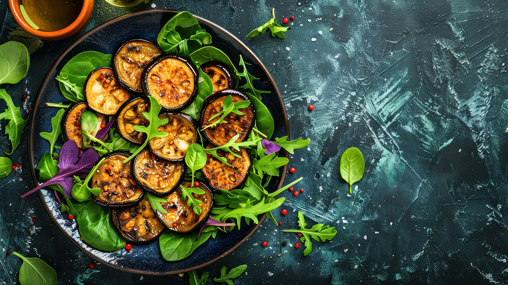 A top-down view of a dark plate filled with grilled eggplant slices and fresh green salad greens, including arugula and spinach, on a dark textured blue-green tabletop.