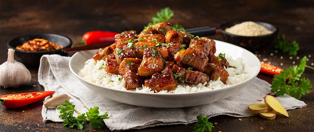 A plated dish of glazed meat over white rice on a dark rustic table, surrounded by garnishes including parsley, garlic, red chili peppers, ginger, and small bowls of seasoning.