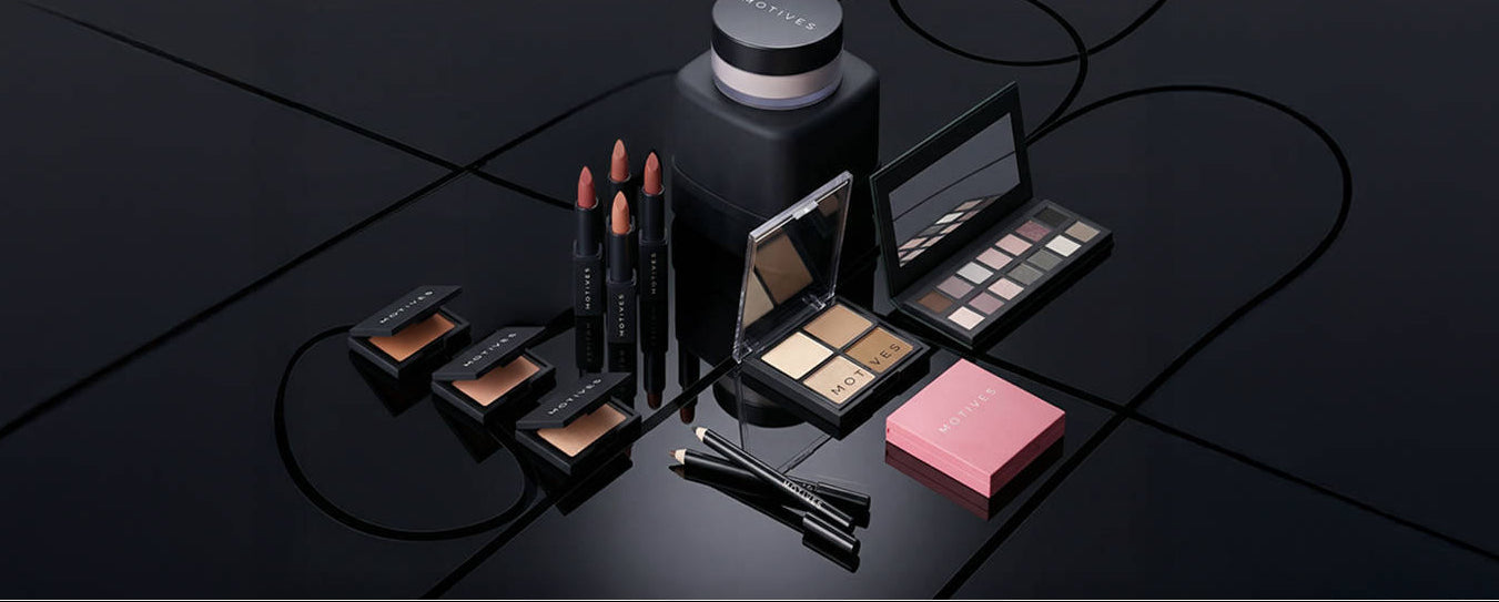 A collection of luxury makeup products from Estée Lauder, including eyeshadows, lipsticks, a compact powder, and a pink compact, arranged on a glossy black surface with elegant curved lines.
