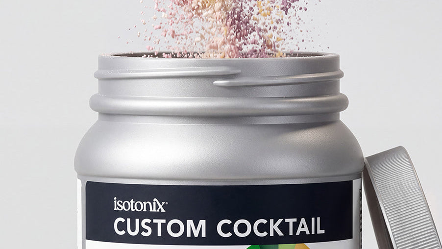 A close-up of an open silver supplement jar labeled 'isotonix CUSTOM COCKTAIL' with colorful powder bursting from the top, and the lid resting beside it on a light gray background.