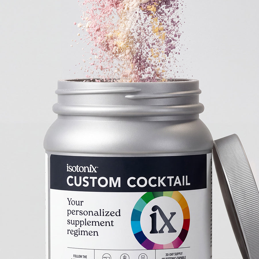 A close-up of an open silver supplement jar labeled 'isotonix CUSTOM COCKTAIL' with colorful powder bursting from the top, and the lid resting beside it on a light gray background.