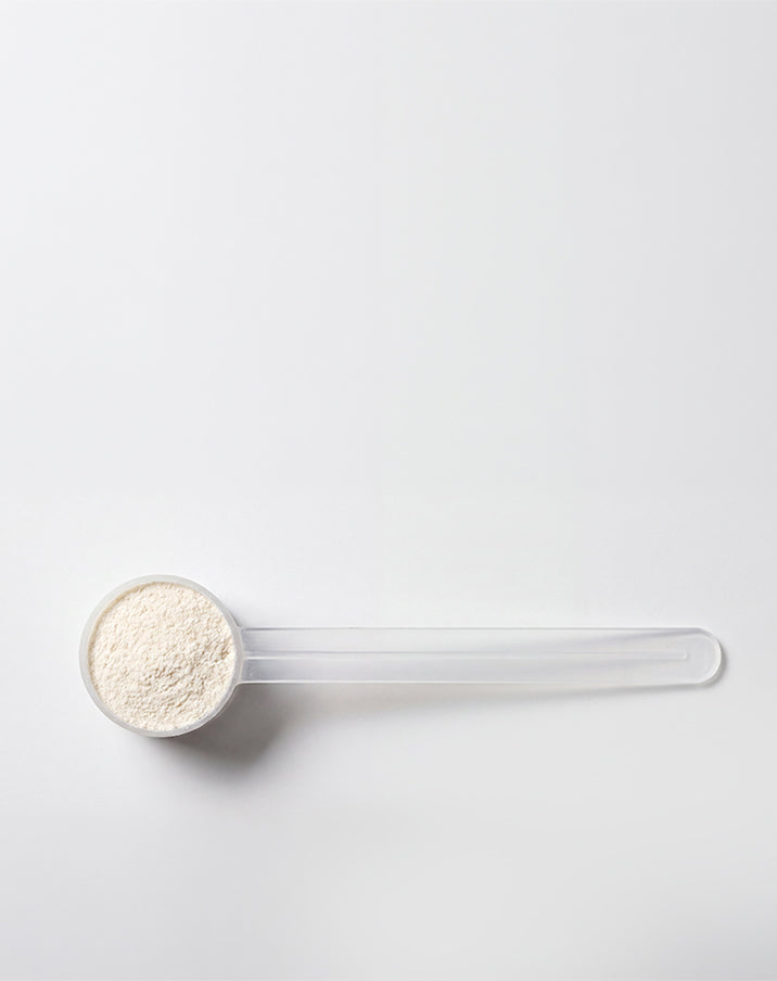 A clear plastic scoop filled with a light beige powder, photographed on a clean white background.