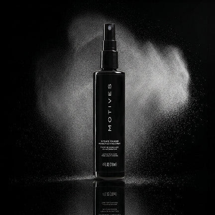 A black spray bottle of MOTIVES Black Powder Hair Fixer standing on a reflective black surface, with white powder visibly spraying from the nozzle against a dark background.