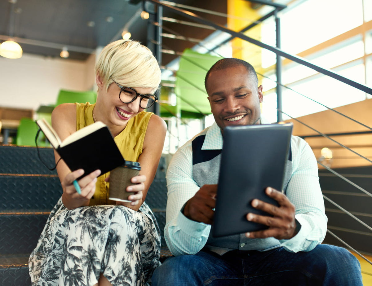 A smiling woman with short blonde hair and glasses holds an open notebook and a coffee cup while sitting next to a man who is looking at a tablet on a staircase in a modern office setting.