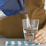 A close-up of a person pouring water from a blue pitcher into a clear glass, with a brown shirt and a blue-and-white striped napkin visible.